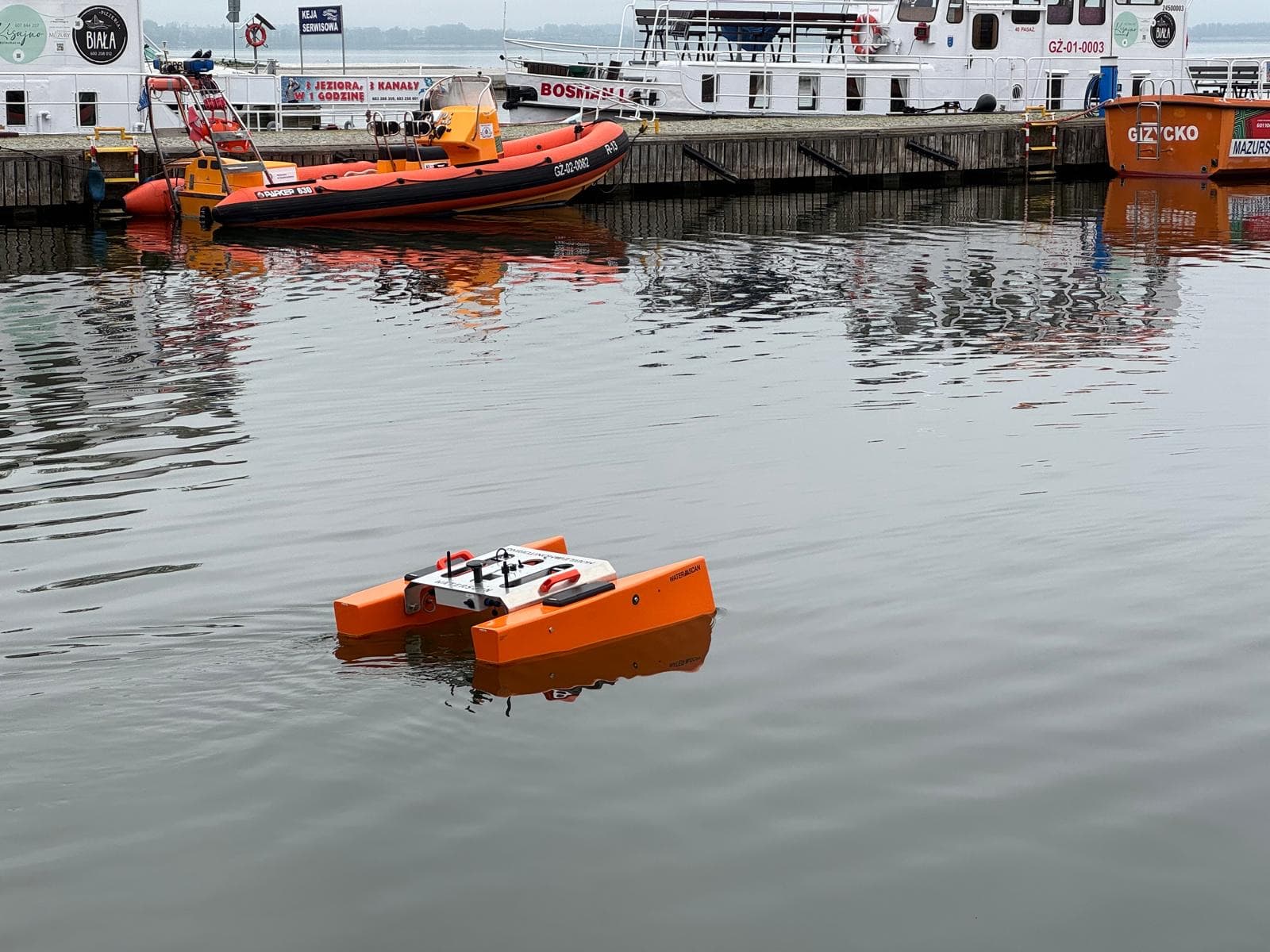 The orange water drone floating in a harbour basin during testing, with boats and ships behind.