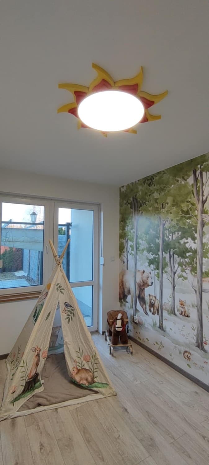 Lampa słońce zamontowana na suficie pokoju dziecięcego z leśną tapetą.